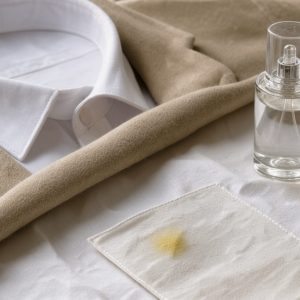 Col de chemise blanche et veste beige protégés par un tissu en coton, avec un vaporisateur de parfum et une légère tache jaune visible sur un échantillon textile.